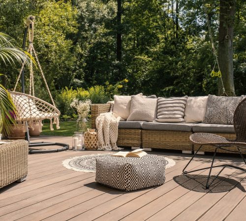 A rattan patio set including a sofa, a table and a chair on a wooden deck in the sunny garden.